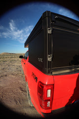 Red pickup with TUNE truck camper, ideal for an RV rental or overland trip. Parked in a dusty desert landscape under blue skies.