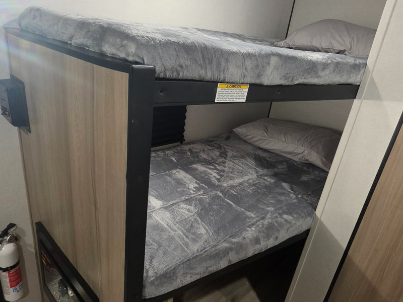  grassy landscape under a clear sky.|RV rental bunk beds feature cozy grey bedding. A safety sticker warns of 300 lbs max capacity. A fire extinguisher is nearby.|RV rental bathroom with a wall-mounted towel rack holding colorful towels — photo 2