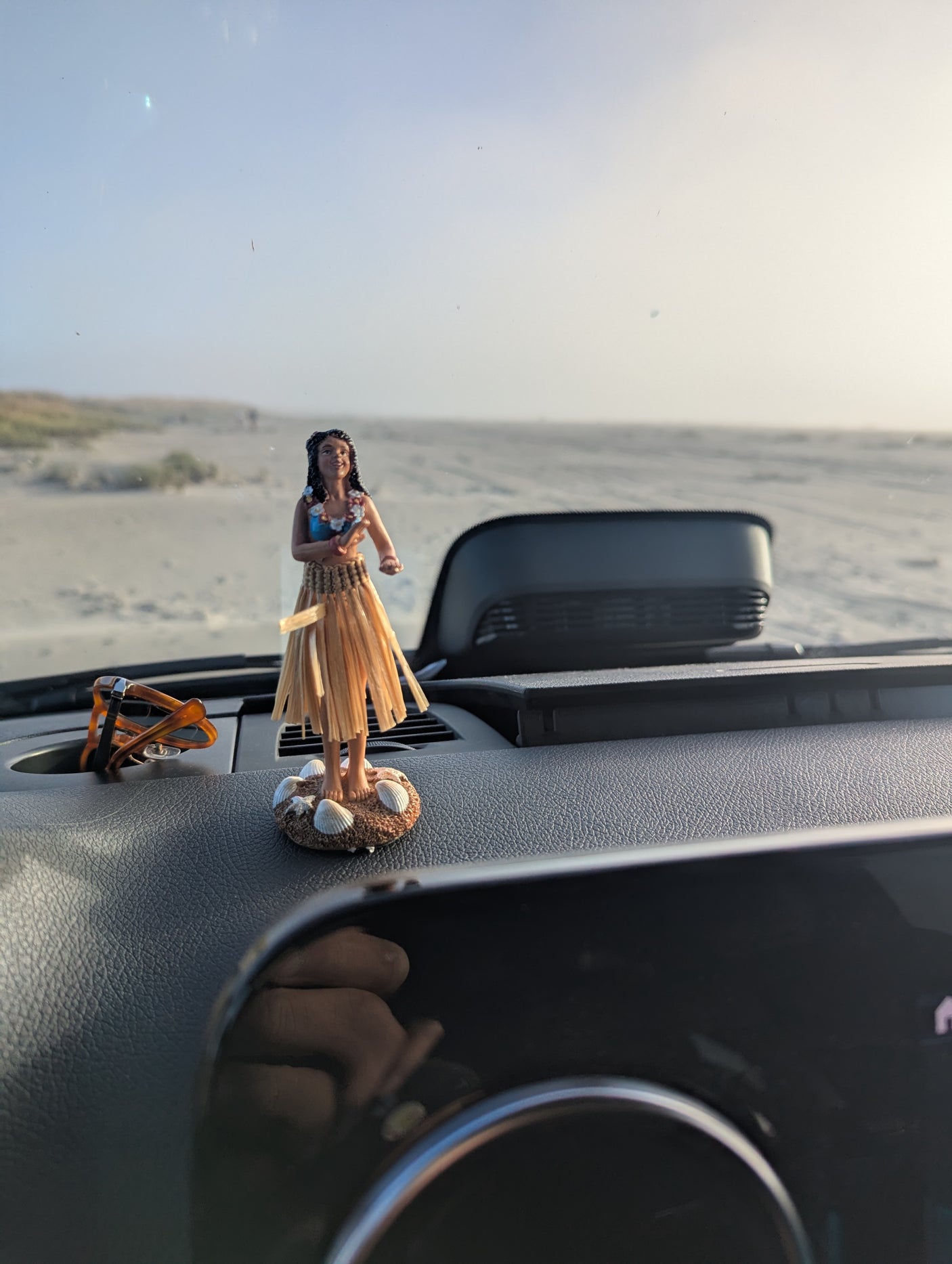 RV rental dashboard scene: a hula girl, sunglasses, and a hand's reflection, with a vast sandy beach beyond.