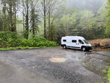 White rv rental parked on a wet gravel lot, surrounded by a misty forest. A person smiles from the driver's seat.