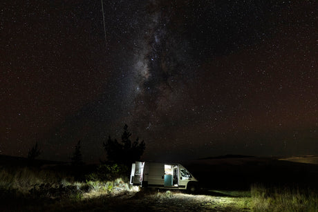 Experience epic stargazing nights with an RV rental. A campervan glows under the Milky Way in a serene, dark landscape.