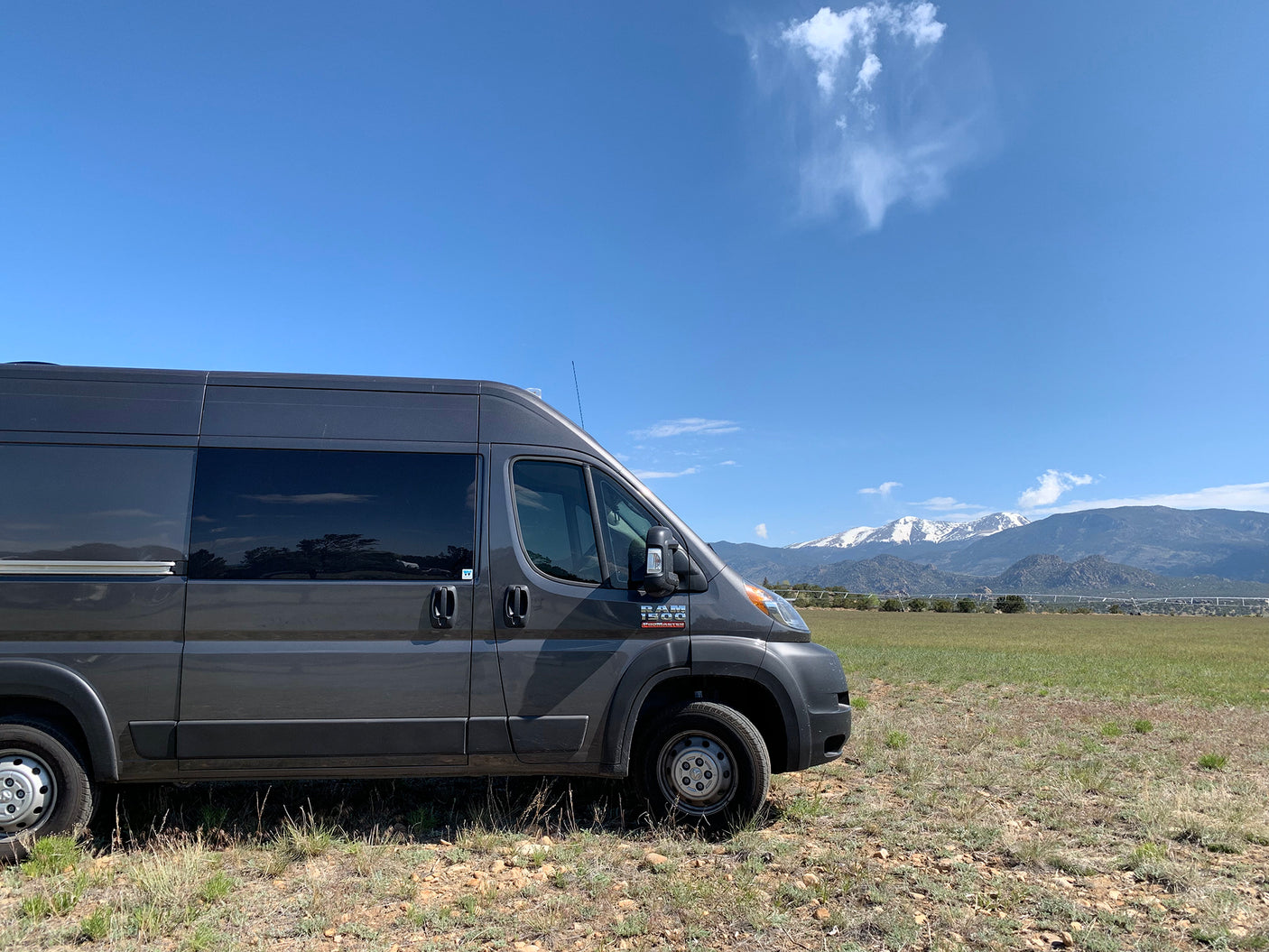 A gray Ram ProMaster RV rental van, parked in a sunny field with distant snow-capped mountains under a blue sky.