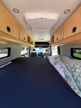 Spacious RV rental interior featuring a comfortable bed, wood overhead cabinets, AC unit, and driver's cabin view.