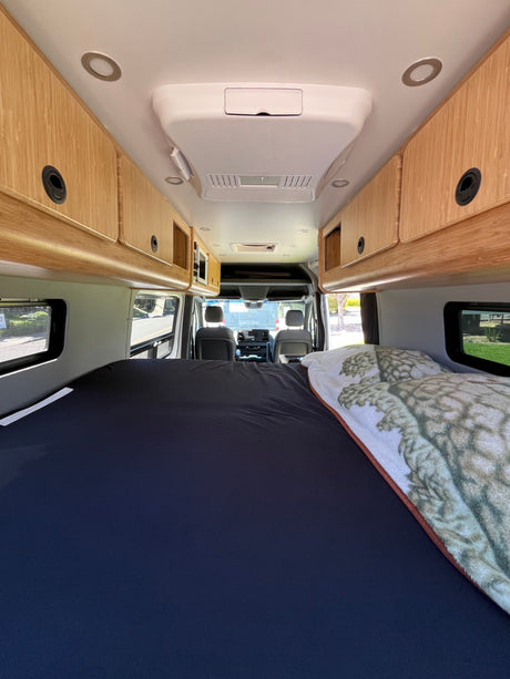 Spacious RV rental interior featuring a comfortable bed, wood overhead cabinets, AC unit, and driver's cabin view.