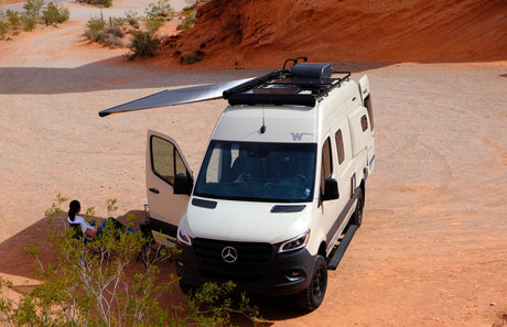 A white Mercedes-Benz rv rental camper van rests in a red desert. Its awning provides shade for a person enjoying the outdoors.