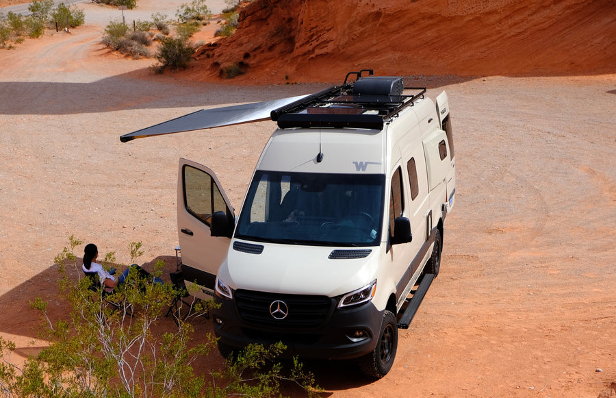 A white Mercedes-Benz rv rental camper van rests in a red desert. Its awning provides shade for a person enjoying the outdoors.