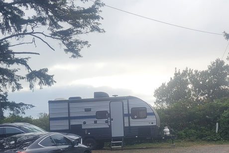 An RV rental travel trailer is parked outdoors under an overcast sky, with cars and trees nearby. Ready for camping!