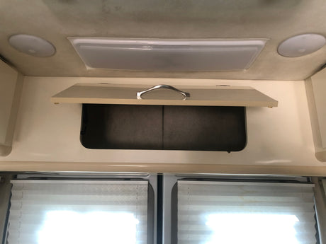 View inside an RV rental showing an open overhead cabinet, two ceiling lights, and pleated window blinds.