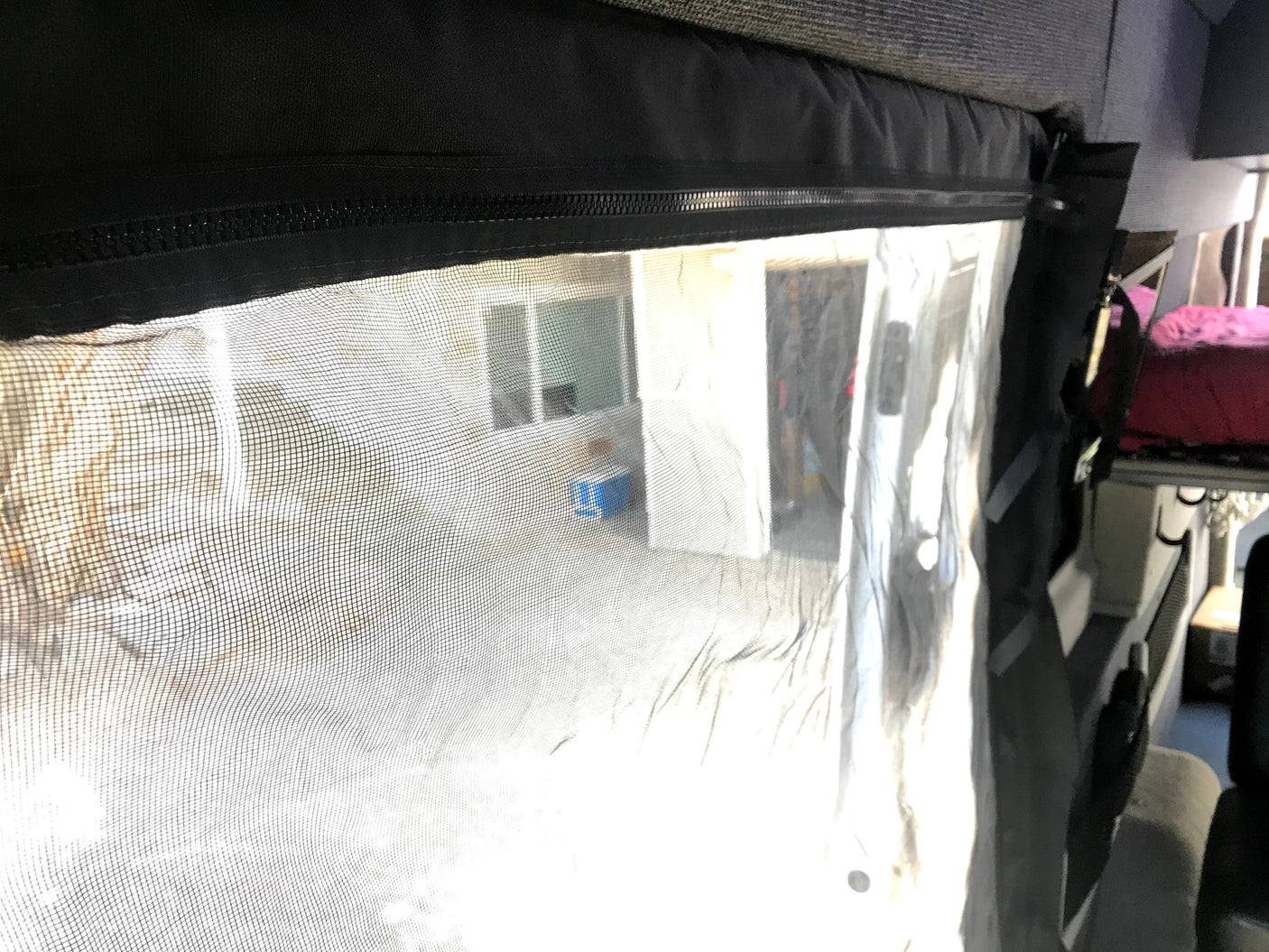 RV rental: View from inside, looking through a zippered mesh screen at an outdoor building & cooler. Interior bed seen right.