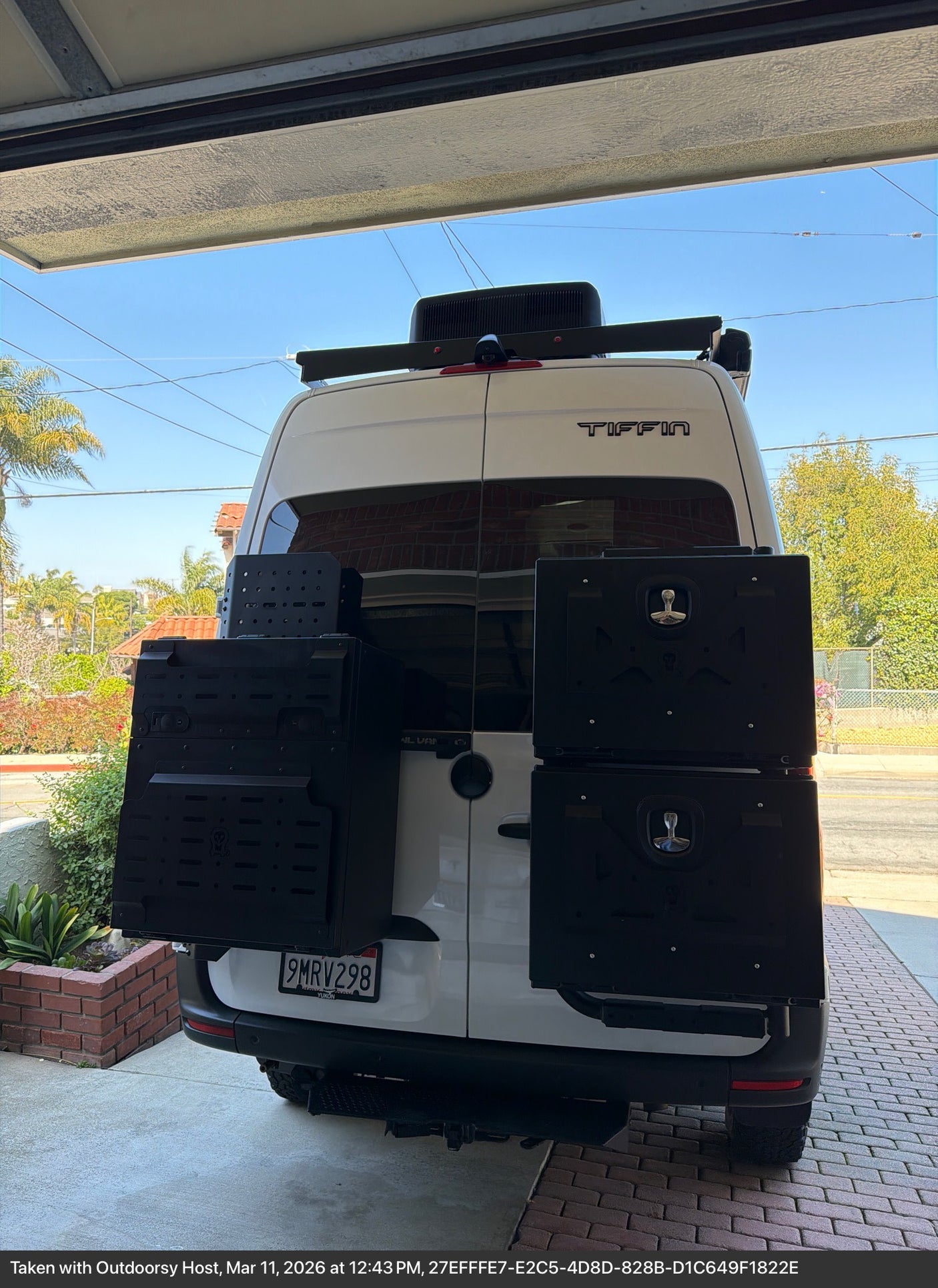 This white Tiffin RV rental van, featuring rear-mounted black storage boxes, is parked under a garage awning.