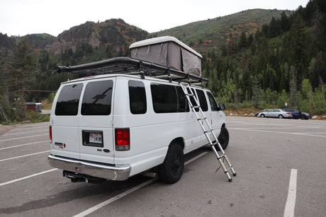 Explore nature with this white rv rental camper van, featuring a rooftop tent & ladder, parked by autumn mountains.