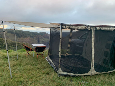 Overland camping setup with a vehicle-attached screen room & awning, chairs, and table, overlooking a scenic valley. Great RV rental alternative!
