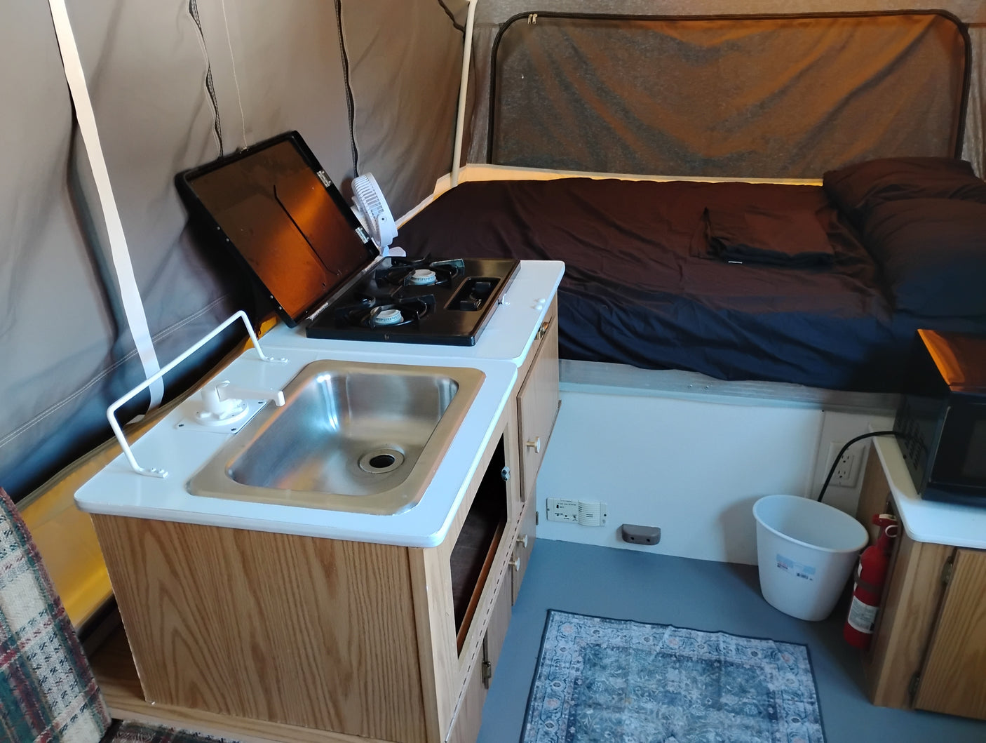 Inside an RV rental pop-up camper: a compact kitchenette with sink, stove, and fan sits opposite a made bed.