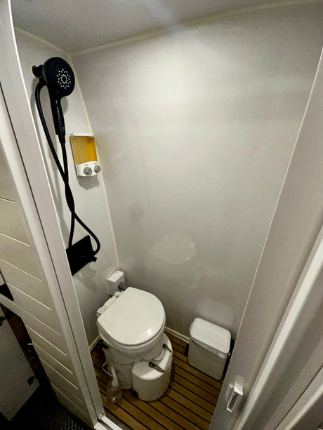 This RV rental bathroom offers a compact design with a composting toilet, shower head, and wall-mounted soap dispenser.
