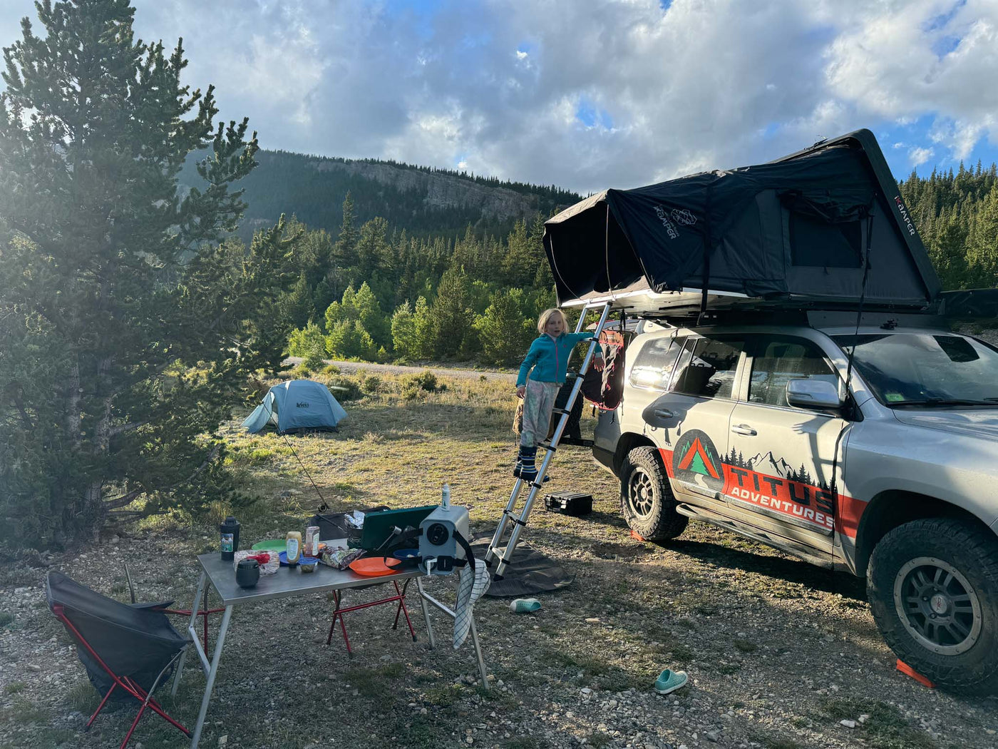  set for camping in a vast desert landscape under blue skies.|RV rental overlanding: Child on ladder to roof-top tent next to camp setup in a scenic outdoor adventure.|An adventure-ready RV rental SUV with rooftop tent drives on a scenic mountain dirt road.|Titus Adventures RV rental — photo 3