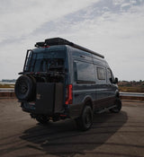 Dark gray Mercedes Sprinter van, an ideal rv rental. Features off-road tires, a ladder, storage box, and roof rack.