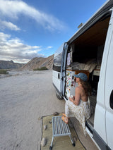 Desert RV rental trip: A woman enjoys coffee from her parked van, embracing remote life amidst mountains.
