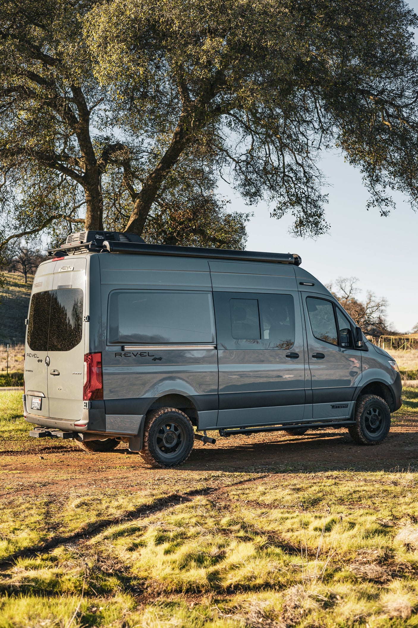 Grey Winnebago Revel 4x4 camper van parked on a sunny dirt path under a tree. Ideal for your next rv rental adventure!