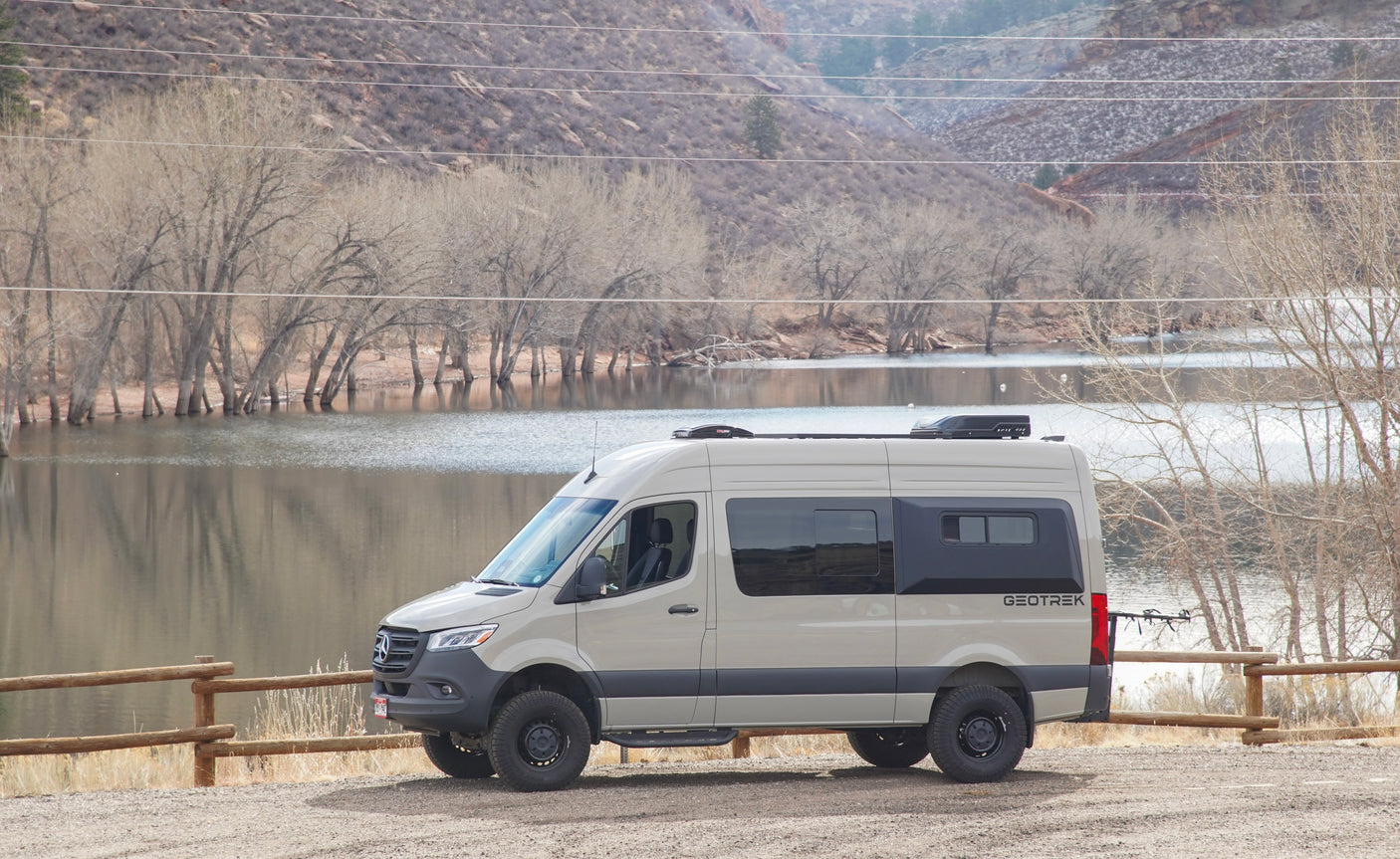 Tan Geotrek RV rental Sprinter van by a lake with bare trees & mountains, ready for outdoor adventure.