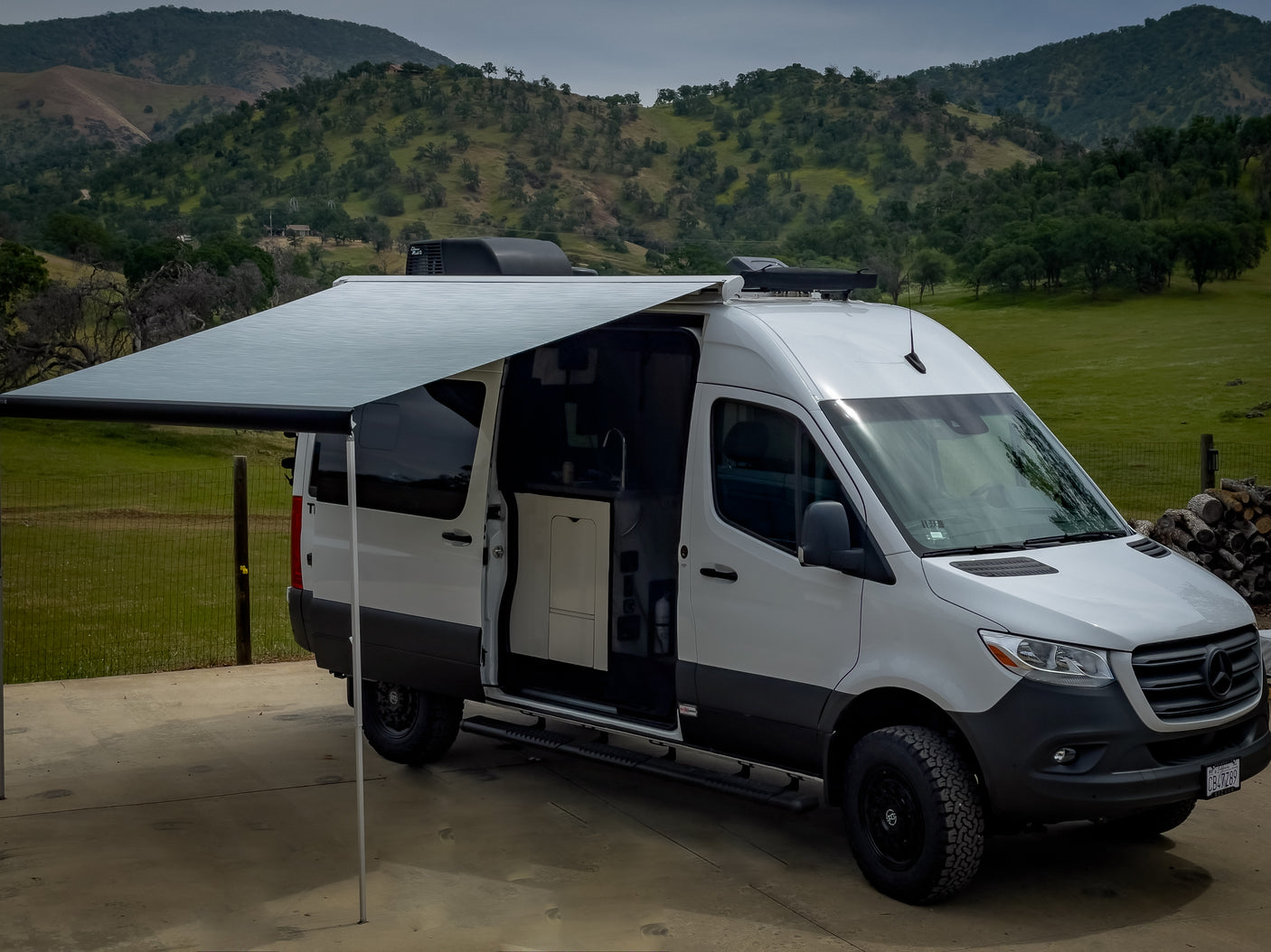 White Sprinter van RV rental with extended awning & open door revealing interior kitchen, parked in scenic hills.