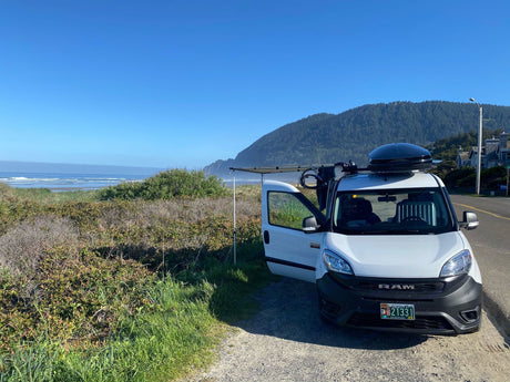 White Ram van, an rv rental, parked by an Oregon beach with an awning out. Mountains and ocean in view under blue sky.