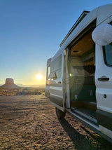 This RV rental offers sunset views over a desert mesa. Its open side door reveals a cozy, wood-paneled interior.