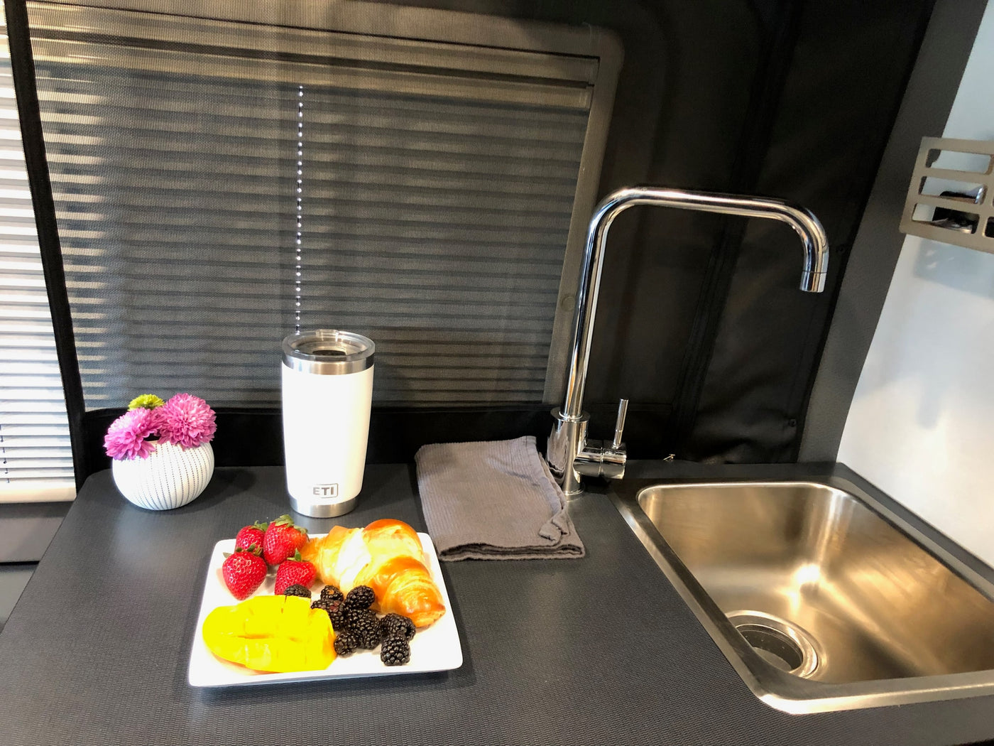 Cozy RV rental interior with a compact kitchen: sink, counter, fresh fruit breakfast, Yeti tumbler, and window blinds. — photo 10