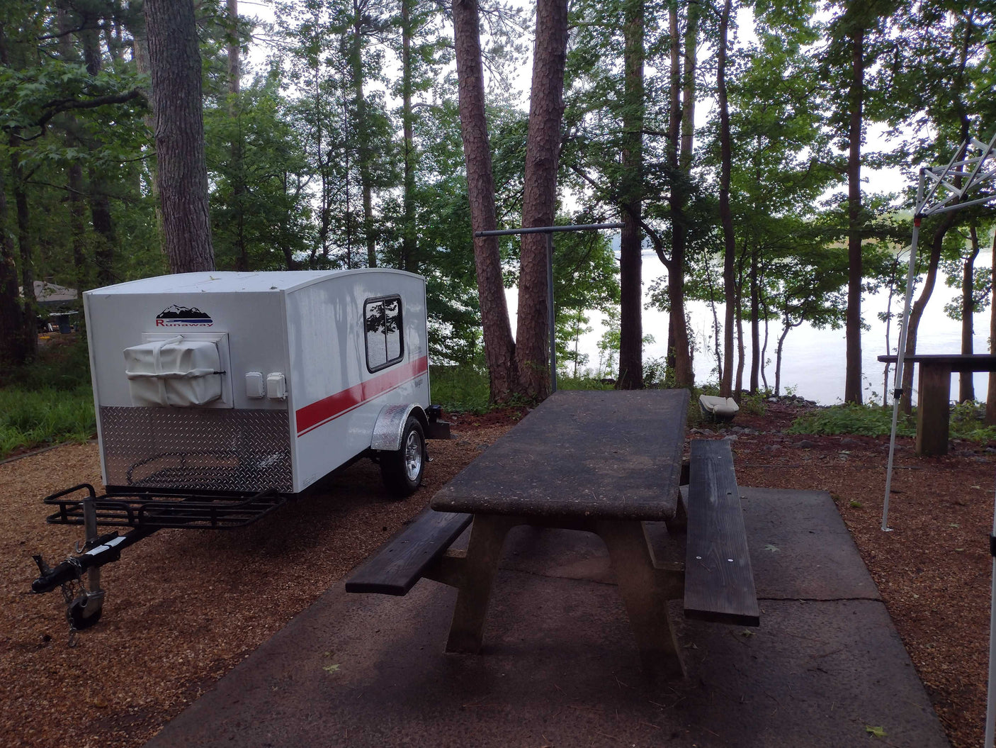 A white Runaway RV rental teardrop camper is parked at a serene lakeside campsite with a picnic table under tall trees.