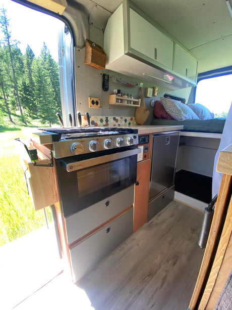 Explore this RV rental's clever camper van interior, featuring a full kitchen with oven, fridge, and a comfy bed, overlooking nature.