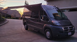 Dark campervan RV rental with extended awning, parked on a driveway at sunset, ready for your next road trip.