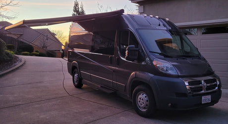 Dark campervan RV rental with extended awning, parked on a driveway at sunset, ready for your next road trip.