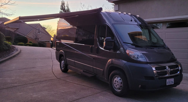 Dark campervan RV rental with extended awning, parked on a driveway at sunset, ready for your next road trip.