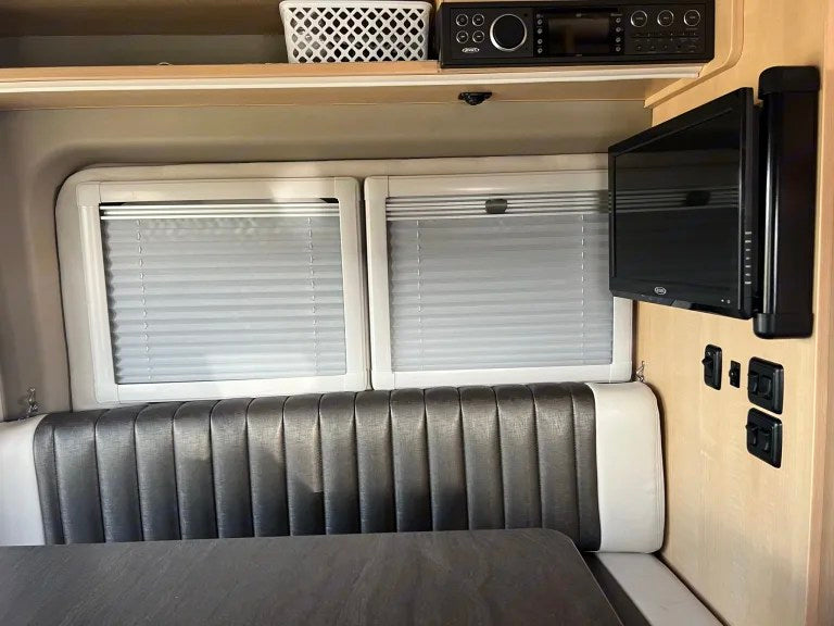 Inside an RV rental: a comfortable gray dinette with white trim, pleated blinds, TV, and entertainment system.