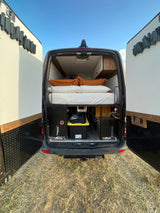 Open rear doors of a dark grey **rv rental** camper van reveal a cozy bed & storage. Parked on dry grass.