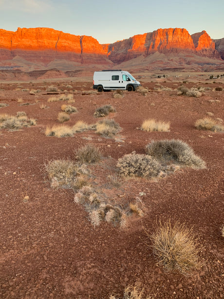 RV rental adventure: White van in a vast red desert, with majestic red rock cliffs glowing under golden hour light.