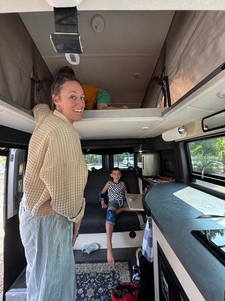 Inside a spacious RV rental, a woman smiles while kids relax on the bunk bed & bench seat, revealing a cozy camper van interior.