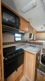 Clean RV rental kitchen with microwave, stove, sink, and wooden cabinets. Rear sleeping area visible.