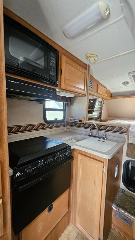 Clean RV rental kitchen with microwave, stove, sink, and wooden cabinets. Rear sleeping area visible.