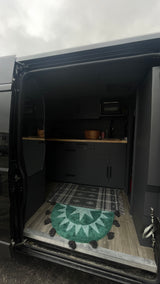 Modern RV rental interior with a compact dark kitchen, wood counters, and patterned rugs seen from the open side door.