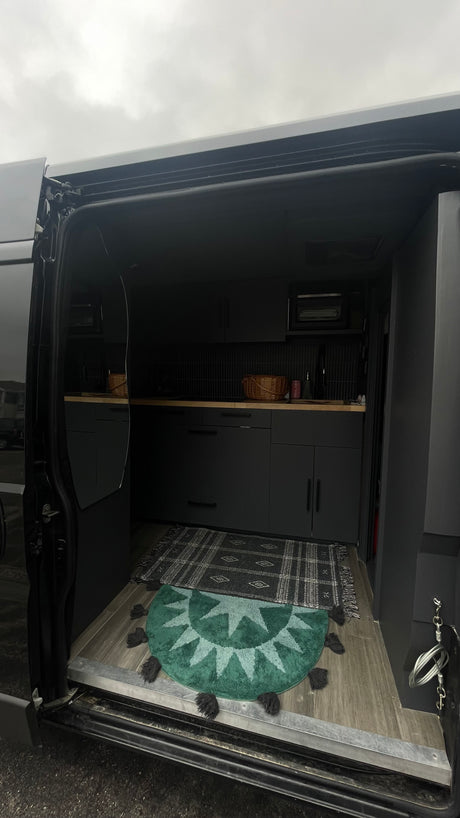 Modern RV rental interior with a compact dark kitchen, wood counters, and patterned rugs seen from the open side door.