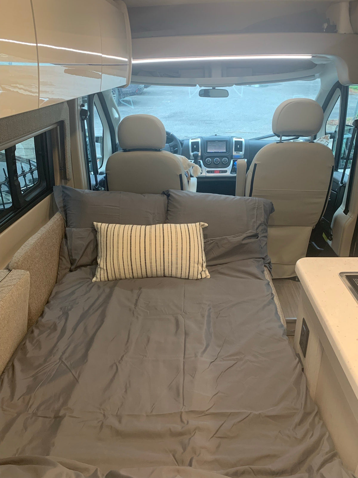 Cozy RV rental interior: Full-size bed with grey bedding, compact kitchen, and front cockpit of a camper van. — photo 16