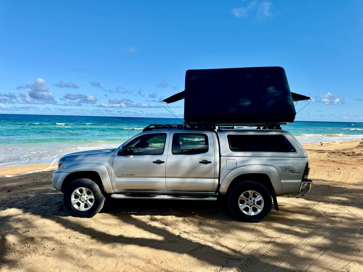 Silver Toyota Tacoma truck with a black rooftop tent, parked on a sandy beach by the blue ocean. Ideal RV rental for off-road camping. — photo 3