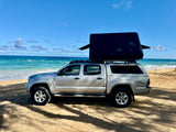 Silver Toyota Tacoma truck with a black rooftop tent, parked on a sandy beach by the blue ocean. Ideal RV rental for off-road camping.
