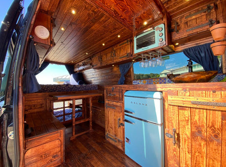 Experience a custom RV rental with a rustic wood interior, featuring retro blue kitchen appliances, cozy bed, and detailed craftsmanship.