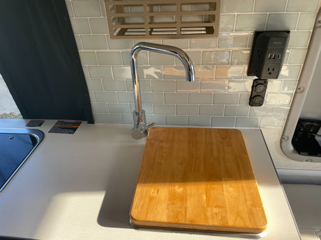 RV rental kitchen: chrome faucet, wooden cutting board on counter. Tiled backsplash with power/USB outlets.