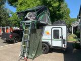 This RV rental, a small travel trailer, features a roof-top tent with ladder & privacy enclosure, parked roadside.