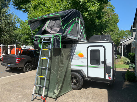 This RV rental, a small travel trailer, features a roof-top tent with ladder & privacy enclosure, parked roadside.