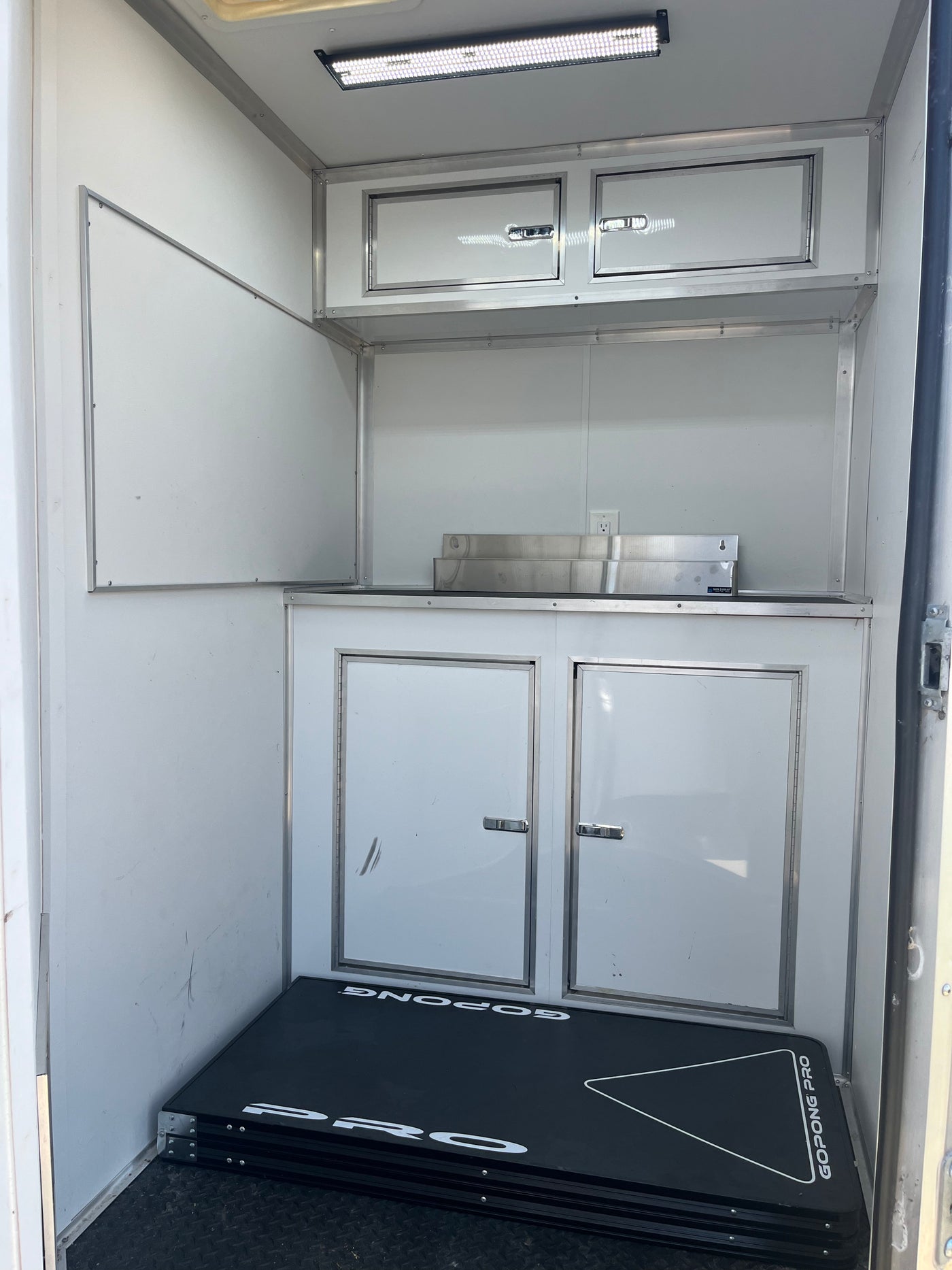 Compact RV rental interior: overhead LED, white cabinets, counter, and a folded GoPong Pro table on the floor. — photo 7
