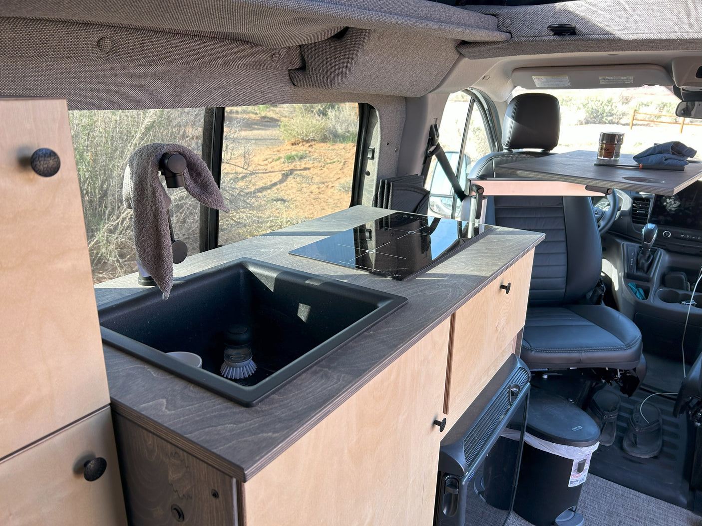 RV rental: Inside a camper van, a compact kitchen with a sink, induction cooktop, and front swivel table. View of desert landscape.