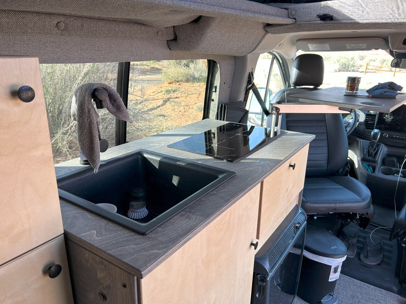 RV rental: Inside a camper van, a compact kitchen with a sink, induction cooktop, and front swivel table. View of desert landscape. — photo 6